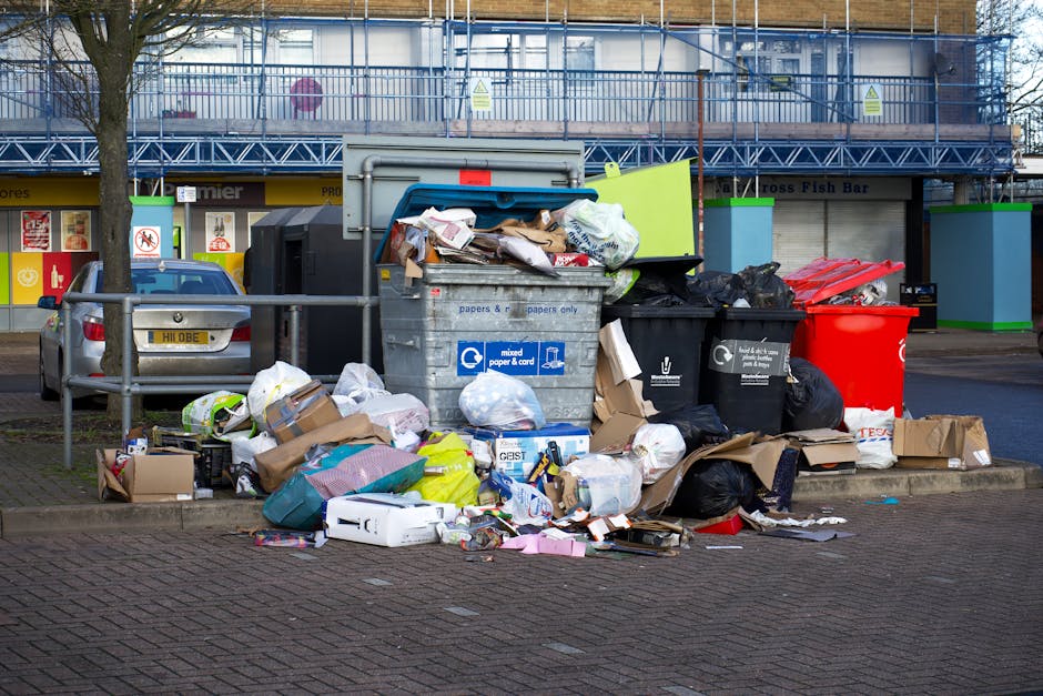 A collection of mixed waste and rubbish is piled around a central metal recycling bin on a city street, with various black, green, and red rubbish bags, cardboard boxes, and loose paper and packaging spilling onto the paved pavement. Behind the waste, there is a silver car parked parallel to the curb, and a row of small retail units with storefronts and signage visible in the background, including one for a fish bar. The central recycling bin is partially open and appears to be overfilled, with paper and cardboard protruding from the top. The scene is outdoors with natural lighting, and a metal railing runs along the edge of the pavement, separating the waste area from the parking space. The surroundings include a small tree on the left, and the background features scaffolding and construction barriers, indicating nearby building work. This image highlights the kind of clutter associated with private or alternative waste collection methods, emphasizing the importance of proper rubbish removal services for maintaining street cleanliness in residential and commercial areas.