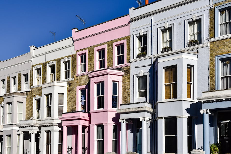 A row of Victorian-style terraced houses on a city street, with facades made of brick and stucco. The buildings feature tall, narrow sash windows with white frames, some adorned with window boxes filled with small plants. One prominent building in the center is painted in pastel pink, with decorative architectural details including protruding bay windows and a cornice at the top. The neighboring houses are finished in white, light blue, and off-white tones, with some brickwork visible beneath the paint. The street is illuminated by natural sunlight, casting shadows that highlight the textures of the facades and creating a clear, bright atmosphere. The image suggests an urban residential area, which could be relevant for independent or private waste collection services related to property or flat clearances in such historic neighborhoods. The visual focus on the building exteriors supports context for services like rubbish removal, especially when considering on-site clearance or alternative waste handling for older city dwellings.