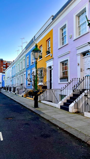 A row of Victorian-style terraced houses on a city street, with facades made of brick and stucco. The buildings feature tall, narrow sash windows with white frames, some adorned with window boxes filled with small plants. One prominent building in the center is painted in pastel pink, with decorative architectural details including protruding bay windows and a cornice at the top. The neighboring houses are finished in white, light blue, and off-white tones, with some brickwork visible beneath the paint. The street is illuminated by natural sunlight, casting shadows that highlight the textures of the facades and creating a clear, bright atmosphere. The image suggests an urban residential area, which could be relevant for independent or private waste collection services related to property or flat clearances in such historic neighborhoods. The visual focus on the building exteriors supports context for services like rubbish removal, especially when considering on-site clearance or alternative waste handling for older city dwellings.