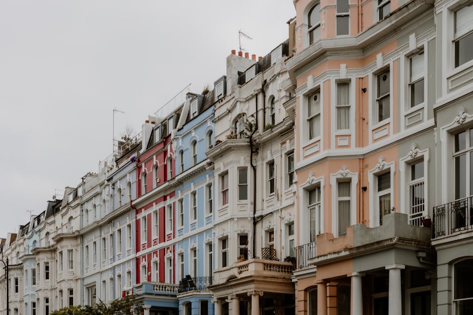 A row of multi-story Victorian-style terraced houses in a residential area, featuring ornate white façade details, large rectangular windows with white frames, and small balconies with wrought iron railings. The buildings are painted in pastel shades including pale pink, white, blue, and cream, with some areas showing decorative stucco mouldings and cornices. The roofs are pitched with slate tiles, chimneys, and antennae visible against a cloudy, overcast sky. The street in front appears to be a regular urban pavement without visible rubbish or debris, suggesting the scene is centered on the architecture and street frontage. The overall environment is calm and orderly, typical of a well-maintained city neighbourhood. The scene's visual context hints at an area where private property maintenance might include external cleaning or minor refuse removal from residential facades, aligning subtly with the concept of independent waste handling and rubbish clearance by services such as those provided by rubbishclearancenottinghill.co.uk, which specializes in rubbish removal for homes and property management in the area.