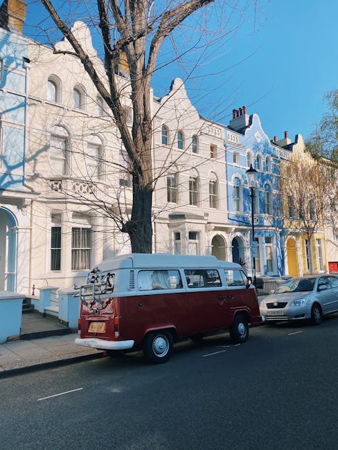 A vintage Volkswagen Type 2 van, painted with a two-tone colour scheme of deep red on the lower body and cream on the upper section, is parked along a residential street in front of a row of Victorian-style terraced houses. The van has a roof rack at the rear loaded with a bicycle, and features classic rounded windows and chrome bumpers, reflecting a retro aesthetic. Behind the van, several modern cars are also parked, including a grey hatchback. The street scene is illuminated by clear daylight with a bright blue sky overhead, and a leafless tree with bare branches extends above the vehicles and houses. The terraced houses exhibit ornate architectural details, with decorative mouldings, sash windows, and a variety of colours including white, blue, and yellow, contributing to a distinctive urban neighbourhood environment. The scene subtly relates to private waste handling and alternative rubbish collection methods by depicting personal vehicles used for transportation and potential rubbish removal activities near residential properties, as exemplified by Rubbish Clearance Notting Hill.