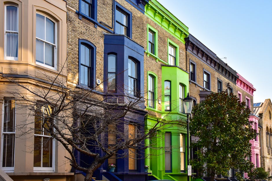 A row of multi-storey residential buildings with ornate brick facades, each painted in distinct vibrant colors including beige, dark blue, bright green, and pink, situated along a city street. The buildings feature large sash windows with white frames, some with decorative molding, and various architectural details such as cornices and stone trim accents. In the foreground, a leafless tree with twisting branches partially obscures the lower sections of the facades, while a lamppost stands near the green building. To the right of the tree, a dense evergreen shrub adds greenery to the scene. The sky above is clear and blue, and the natural daylight highlights the colors and textures of the building exteriors. This urban streetscape exemplifies the attention to detail typical of city architecture and may relate to private property clearance or building refurbishments by companies like rubbishclearancenottinghill.co.uk, focusing on the removal of waste or debris associated with property maintenance or refurbishment projects.