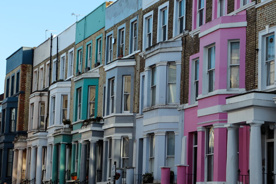 A row of Victorian terraced houses in Notting Hill, characterized by their colorful facades and bay windows. The buildings feature a variety of pastel shades, including blue, white, turquoise, and pink, with decorative white trim and molding accents around the windows and doors. The brickwork on some of the upper sections is visible, contrasting with the painted surfaces. Several of the houses have small front gardens or steps leading up to the entrances, with some potted plants and small garden ornaments visible. The sky above is clear, highlighting the architectural details and vibrant coloring of the facades. This scene reflects typical residential architecture in London’s Notting Hill area, which can be complemented or maintained by local rubbish removal or waste clearance services, especially during property renovations or deep cleans before lettings.