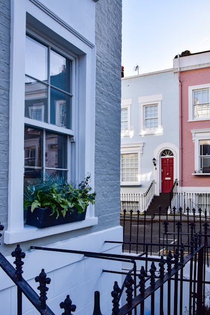 A row of multi-storey residential buildings with ornate brick facades, each painted in distinct vibrant colors including beige, dark blue, bright green, and pink, situated along a city street. The buildings feature large sash windows with white frames, some with decorative molding, and various architectural details such as cornices and stone trim accents. In the foreground, a leafless tree with twisting branches partially obscures the lower sections of the facades, while a lamppost stands near the green building. To the right of the tree, a dense evergreen shrub adds greenery to the scene. The sky above is clear and blue, and the natural daylight highlights the colors and textures of the building exteriors. This urban streetscape exemplifies the attention to detail typical of city architecture and may relate to private property clearance or building refurbishments by companies like rubbishclearancenottinghill.co.uk, focusing on the removal of waste or debris associated with property maintenance or refurbishment projects.
