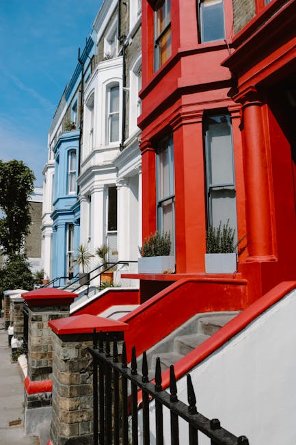 The image displays a row of Victorian-style terraced houses on a residential street, with the building on the right painted in a bold red colour and the adjacent house on the left in a light blue shade. The red house has large sash windows with black frames, and beneath the windows are white window sills. The houses feature bay windows on the ground and first floors, with decorative architectural details such as pillar-like supports at the front corners of the red building. In front of these houses, stone steps with metallic railings lead up to black-painted front doors, each guarded by small brick and stone boundary walls topped with black iron fencing. The steps of the red house are partially visible, with the sidewalk and pavement nearby. The scene is well-lit, indicating bright daylight, with a clear blue sky and some greenery, including small plants in window boxes and a few trees visible in the background. This exterior setting exemplifies typical urban residential architecture, where private properties are maintained with distinct colour schemes, and the scene subtly relates to the need for periodic external cleaning or maintenance, often associated with property management companies such as those involved in waste or rubbish clearance.