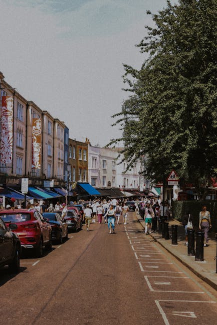 A busy street scene in Notting Hill featuring a row of multi-story buildings with colorful facades, some with shopfronts and awnings on the ground level. The road has parked cars on both sides and a central lane designated for vehicle movement, with a series of parking bays marked along the curb. Pedestrians are walking along the pavement and crossing the street, some carrying shopping bags or engaging in conversation. Large leafy trees line the right side of the street, providing shade and greenery, while the buildings on the left are painted in pastel shades of pink, blue, yellow, and white, with textured exteriors showing signs of weathering. The sky above is overcast, casting a diffuse light over the scene, giving it a vibrant yet muted atmosphere typical of urban areas with active local markets or shops. Occasionally, the scene includes visible signs and street furniture like bollards and traffic signs, with some shop awnings extending outward, suggesting commercial activity and a lively pedestrian environment relevant to private waste collection or rubbish removal services in inner-city areas like Notting Hill.