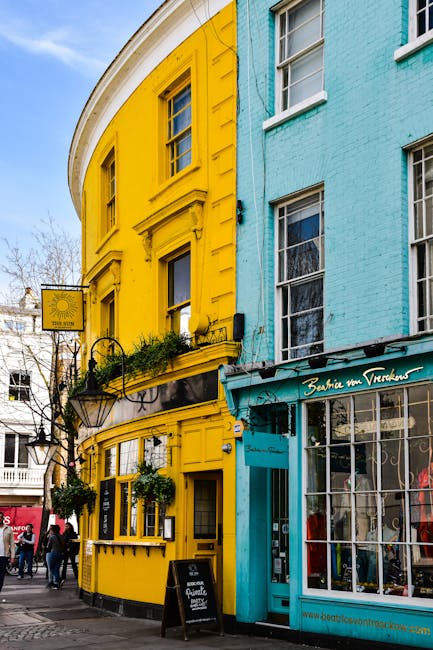 A street scene featuring a brightly painted, curved corner building with a vibrant yellow façade and decorative architectural details such as window surrounds and a small balcony with hanging plants. Adjacent to it is a light blue-painted building with large window panels and a shopfront displaying items behind glass. A signboard made of black metal with a glass lantern-style fixture is mounted on the yellow building, promoting a café or local business. In the foreground, pedestrians are walking along the pavement, and there is a small blackboard sign placed on the sidewalk for advertising. The environment appears to be a lively urban area with a mix of commercial and retail spaces, and the clear sky and daylight enhance the vivid colours of the buildings. This scene may reflect the context of private or independent waste management or disposal typical of a busy shopping or dining district, with potential for rubbish clearance or collection services to operate in such environments, as seen in professional waste removal contexts handled by companies like rubbishclearancenottinghill.co.uk.