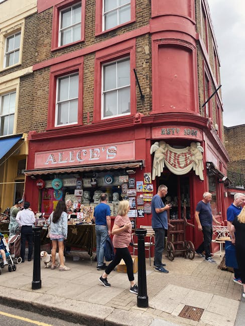 A busy street scene in front of a red-painted building with a vintage appearance, featuring a storefront named 'ALICE'S' established in 1966. The building has large windows with white frames on the upper floors, and the ground level features outdoor displays of various decorative items and signs attached to the facade. People are gathered around the shop, some browsing the items on display, while others are walking past or standing and chatting. A woman wearing a pink top and black trousers is walking, and a man in a blue shirt is checking his phone near the entrance. Several individuals are seen with casual attire, and one person is pushing a stroller with a dog walking nearby. The alleyway area is bordered by black bollards, and the overall scene suggests a lively market or shopping area associated with private collections or independent vendors, aligning with alternative waste handling or rubbish removal services that often operate outside of standard municipal collection systems.