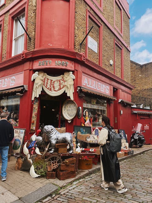 A street scene featuring a brightly painted, curved corner building with a vibrant yellow façade and decorative architectural details such as window surrounds and a small balcony with hanging plants. Adjacent to it is a light blue-painted building with large window panels and a shopfront displaying items behind glass. A signboard made of black metal with a glass lantern-style fixture is mounted on the yellow building, promoting a café or local business. In the foreground, pedestrians are walking along the pavement, and there is a small blackboard sign placed on the sidewalk for advertising. The environment appears to be a lively urban area with a mix of commercial and retail spaces, and the clear sky and daylight enhance the vivid colours of the buildings. This scene may reflect the context of private or independent waste management or disposal typical of a busy shopping or dining district, with potential for rubbish clearance or collection services to operate in such environments, as seen in professional waste removal contexts handled by companies like rubbishclearancenottinghill.co.uk.