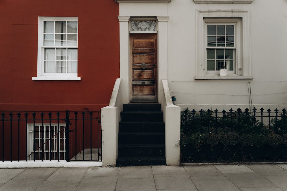 The image depicts a residential building with a central set of black wooden stairs leading up to an aged, weathered wooden front door positioned between two different coloured facades—red on the left and white on the right. The stairs have white-painted concrete side walls, and the door features a dark metal door handle with a small window above, partially covered by decorative frosted glass. To the left of the stairs, a short black metal fence runs parallel to the pavement, enclosing a small garden bed with low shrubs and plants. The red section of the building has a double-hung white window with sash bars on the upper and lower panes, while the white section features a similar double-hung window with a white frame, placed above the stairs, with a small white pot on the windowsill. The setting appears to be an urban street, possibly in a quiet neighbourhood, with natural daylight illuminating the scene. The image relates to private property access and the maintenance of residential exteriors, often associated with services like rubbish clearance or on-site waste removal, as exemplified by companies like rubbishclearancenottinghill.co.uk.