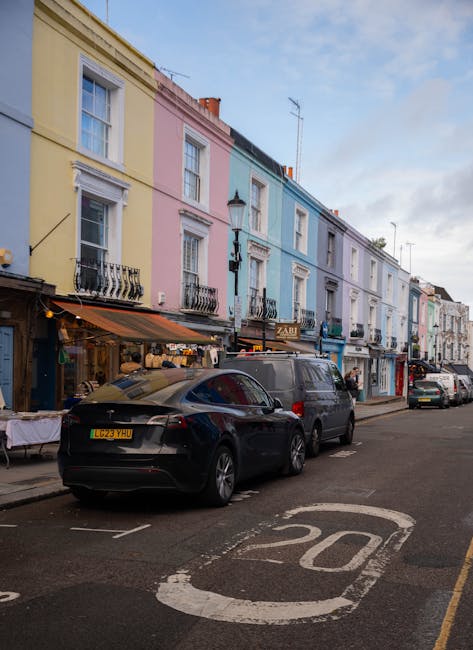 A row of closely positioned two to three-storey terraced buildings painted in pastel shades of yellow, pink, light blue, and white, lining a narrow street. The ground floors of these buildings feature small shops and cafes, with signs visible above the entrances. In the foreground, several parked cars, including a black modern hatchback and a grey van, are aligned along the curb. The street has a marked speed limit of 20 miles per hour painted on the asphalt. Above, the sky is partly cloudy with patches of blue, and a street lamp is mounted on the light blue building. The scene captures an urban street typical of a residential and commercial area, reflecting a setting where private rubbish collection and waste clearance services, such as those offered by rubbishclearancenottinghill.co.uk, could be relevant for domestic or small business waste management needs.