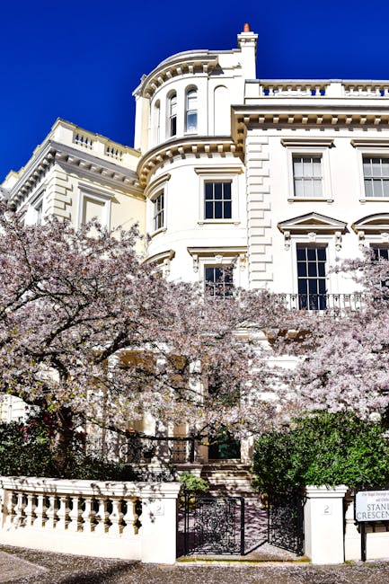 A large, elegant white Victorian-style building with intricate architectural details, including rounded bay windows and decorative cornices, set against a bright blue sky. In the foreground, a flowering cherry tree with pale pink blossoms partially obscures the lower part of the building, while lush green shrubs and a stone retaining wall with a white balustrade run along the front. A black iron gate and steps lead up to the entrance, and a small street sign marks the street name nearby. The scene showcases a well-maintained residential property, characteristic of upscale neighbourhoods, with no visible rubbish or waste, though the setting aligns with private property often associated with independent rubbish collection or on-site clearance in local areas.