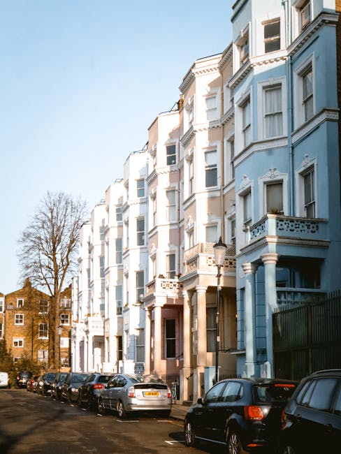 A row of closely positioned two to three-storey terraced buildings painted in pastel shades of yellow, pink, light blue, and white, lining a narrow street. The ground floors of these buildings feature small shops and cafes, with signs visible above the entrances. In the foreground, several parked cars, including a black modern hatchback and a grey van, are aligned along the curb. The street has a marked speed limit of 20 miles per hour painted on the asphalt. Above, the sky is partly cloudy with patches of blue, and a street lamp is mounted on the light blue building. The scene captures an urban street typical of a residential and commercial area, reflecting a setting where private rubbish collection and waste clearance services, such as those offered by rubbishclearancenottinghill.co.uk, could be relevant for domestic or small business waste management needs.